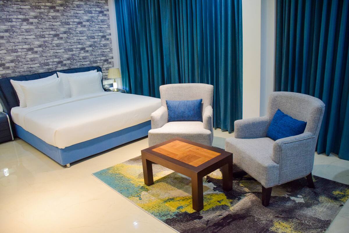 A modern hotel room with a large bed, two armchairs, a wooden coffee table, and a patterned rug.