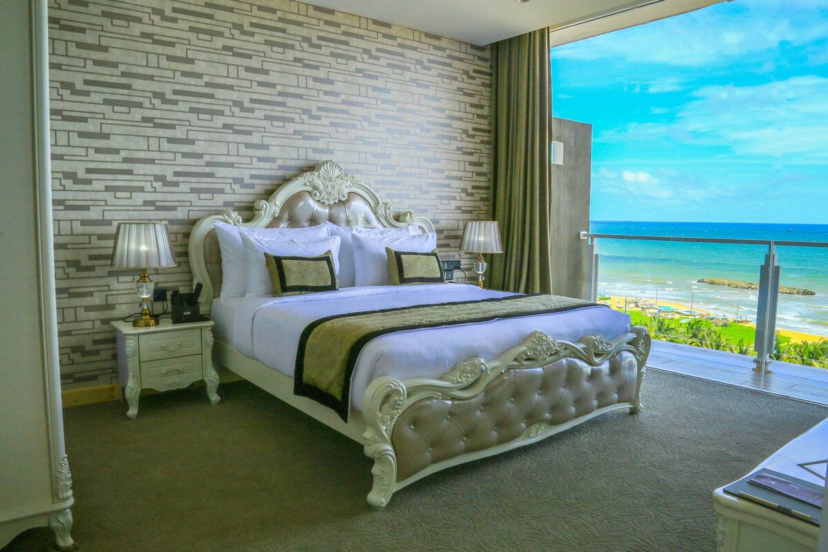 A large bed with a tufted headboard, two bedside tables with lamps, and a view of the ocean through a balcony.