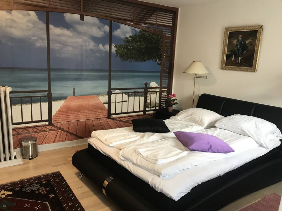 Un dormitorio con cama, ventana con vista al mar, lámpara y un cuadro en la pared.