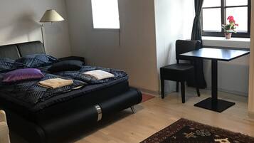 Quarto com cama, escrivaninha, cadeira, janela com cortinas, pequena mesa com planta em vaso e tapete estampado.