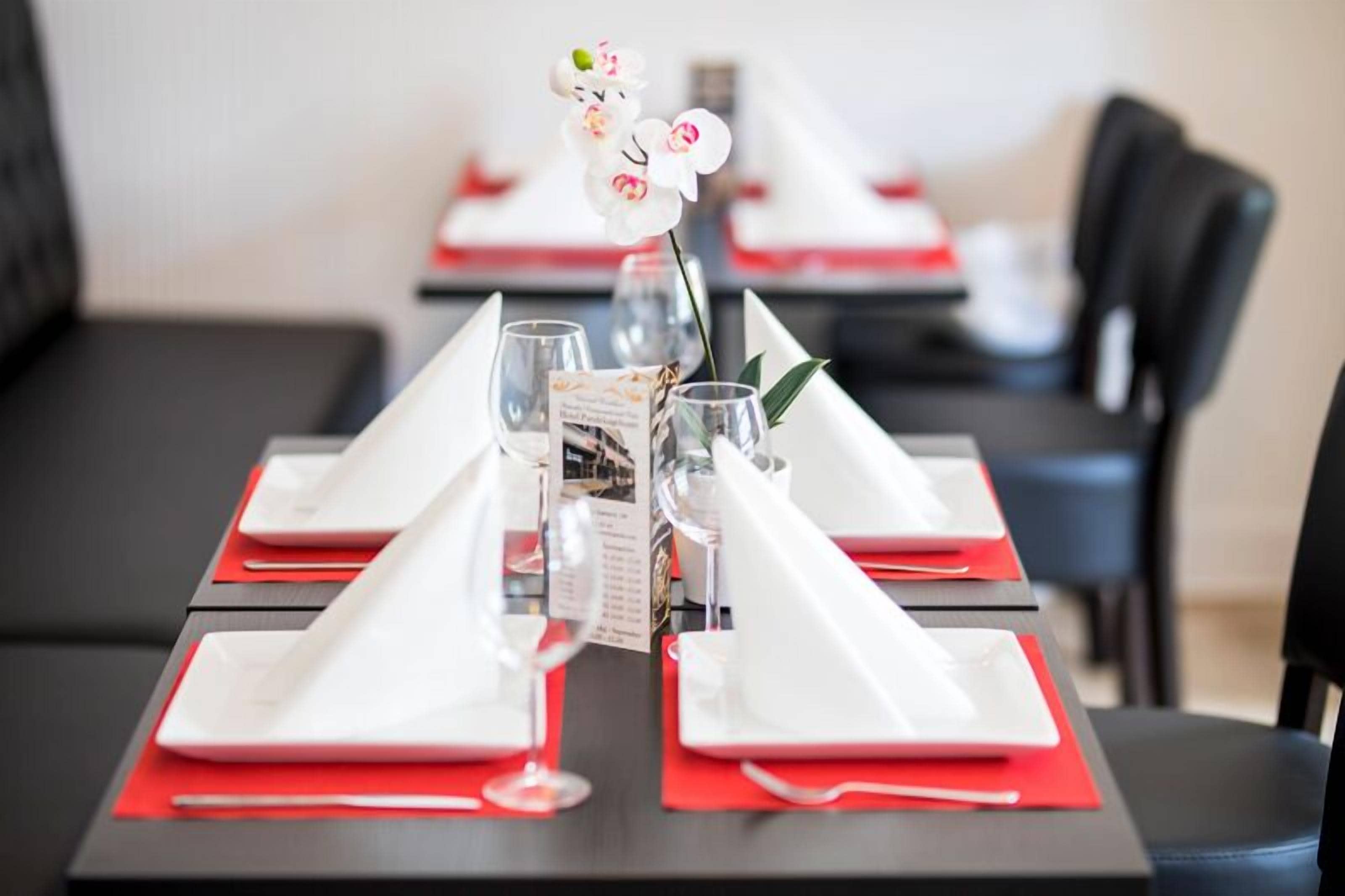 Une table à manger dressée avec de l’argenterie blanche, des serviettes rouges et des verres transparents, accompagnée d’un menu et d’une orchidée blanche.