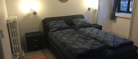 Un dormitorio con cama, mesita de noche, radiador y ventana con cortinas.