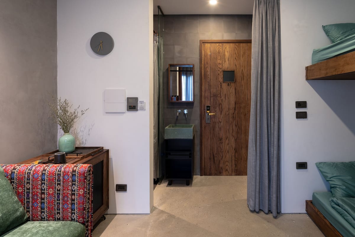 A room with a bed, a small table with a vase, a clock, a wooden door, a sink, and a mirror.