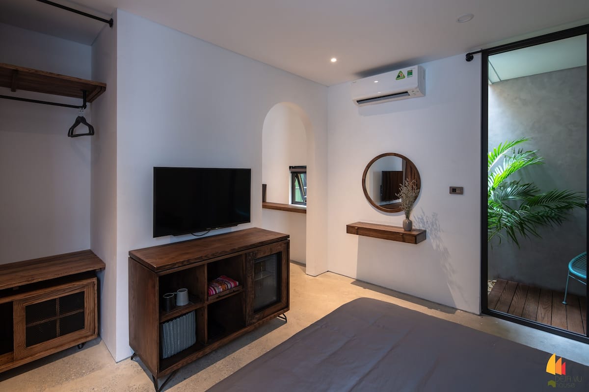 A modern living room with a flat-screen TV, wooden furniture, a round mirror, and a sliding glass door leading to an outdoor area with a plant.