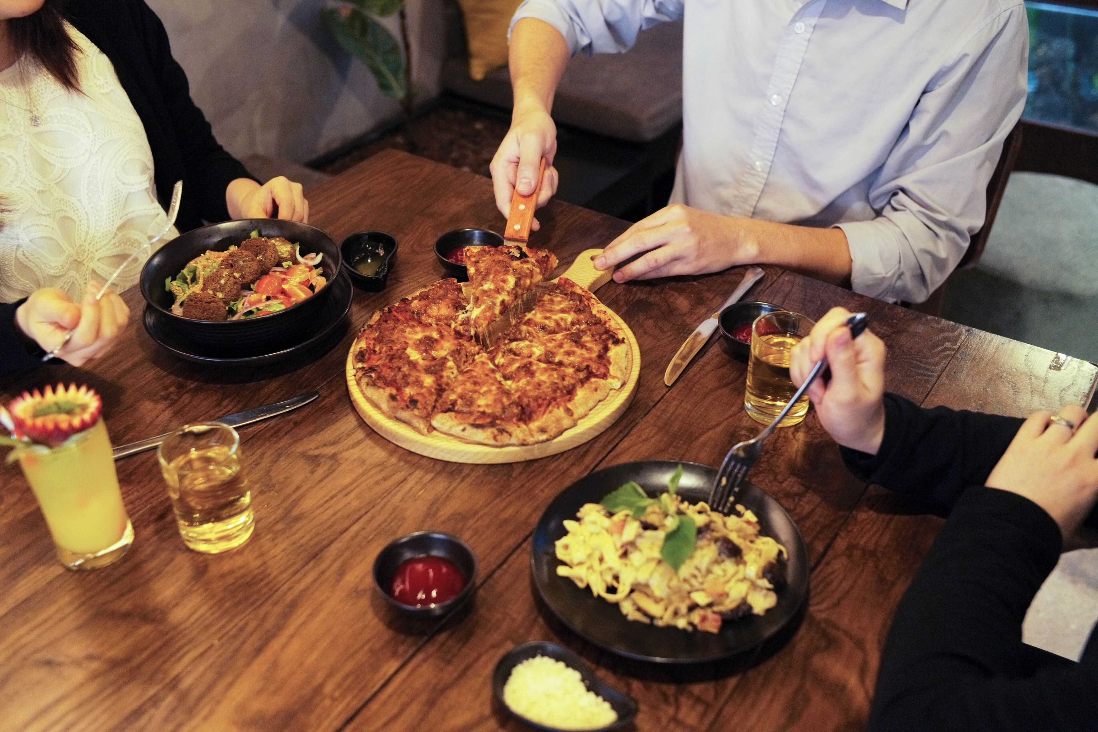 A group of people dining at a wooden table with various dishes, including pizza and pasta, and drinks.