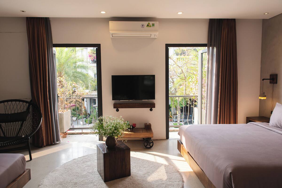A hotel room with a bed, a TV mounted on the wall, a small table with a plant, and a view of the outdoors through large windows.