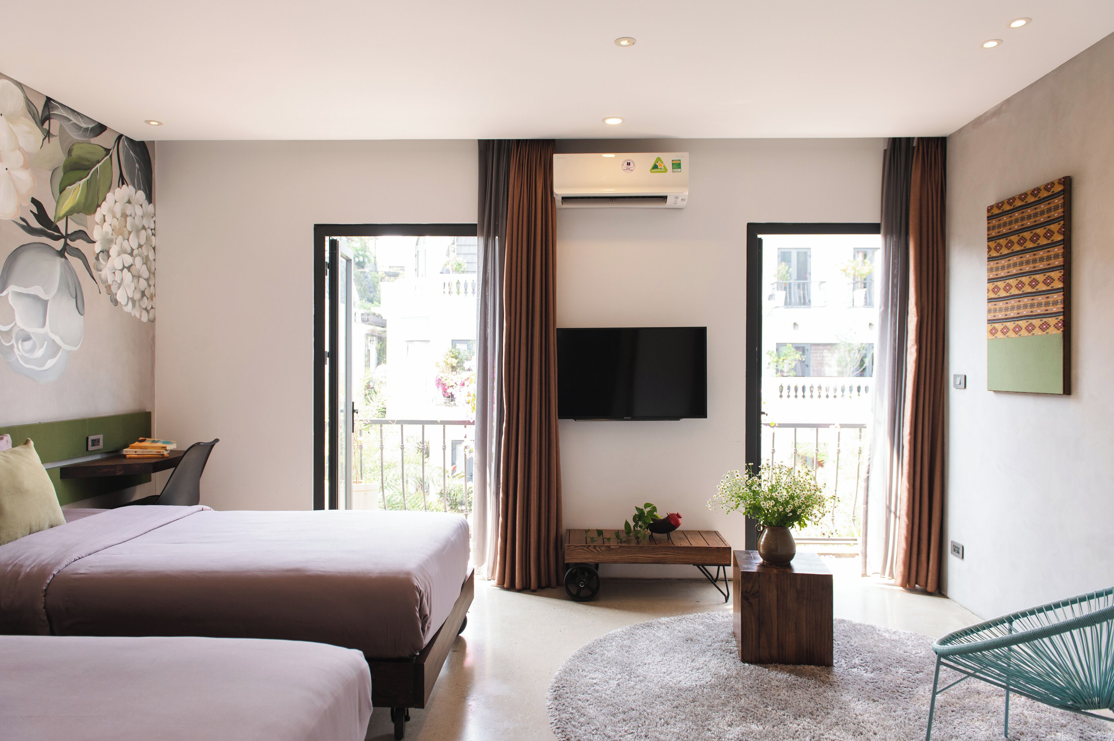 A hotel room with two beds, a flat-screen TV, a desk with a chair, a small table with a plant, and a view of the outside balcony.