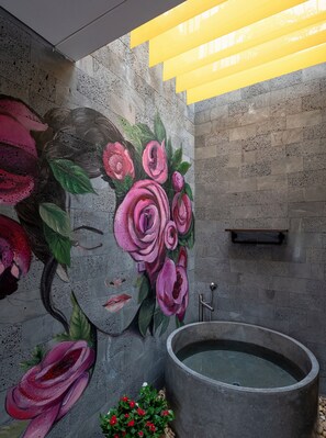 Un baño con una bañera de piedra grande, un mural en la pared de una mujer con rosas, y un accesorio de luz amarilla en el techo.