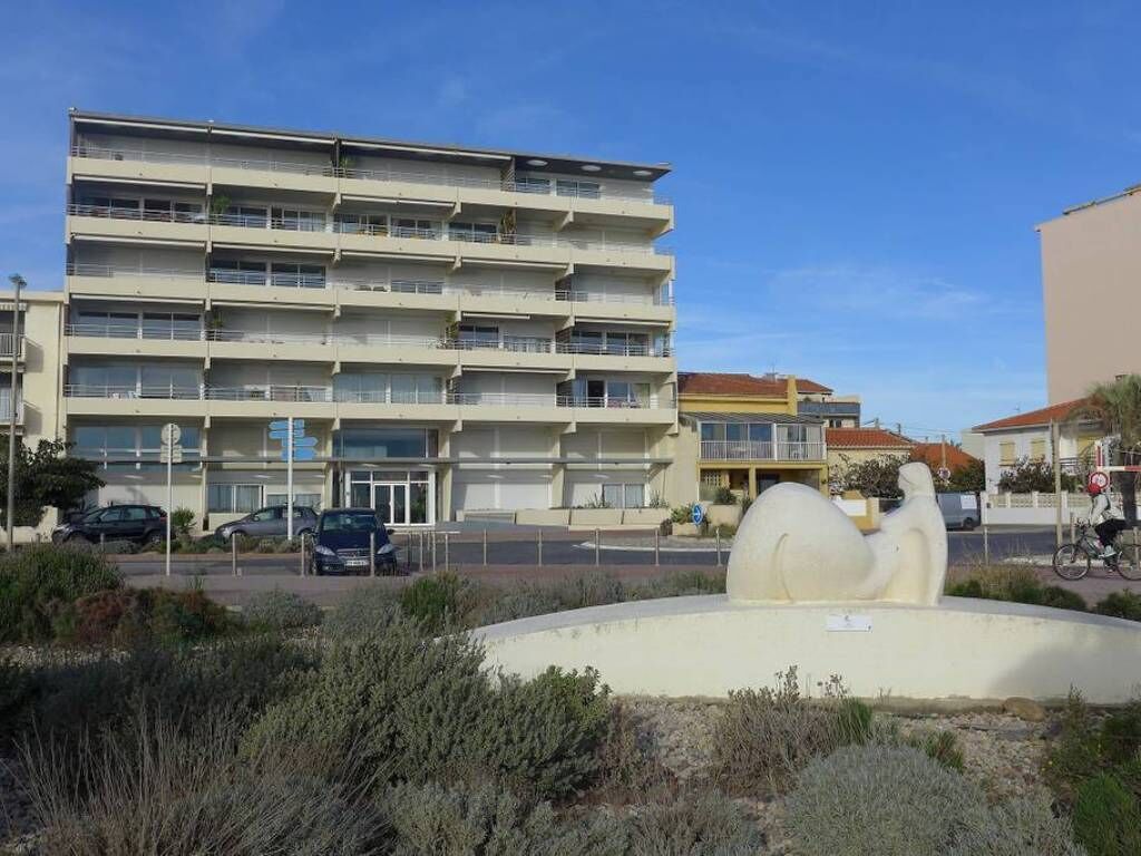 A multi-story building with balconies, a parking area, and a sculpture in the foreground.