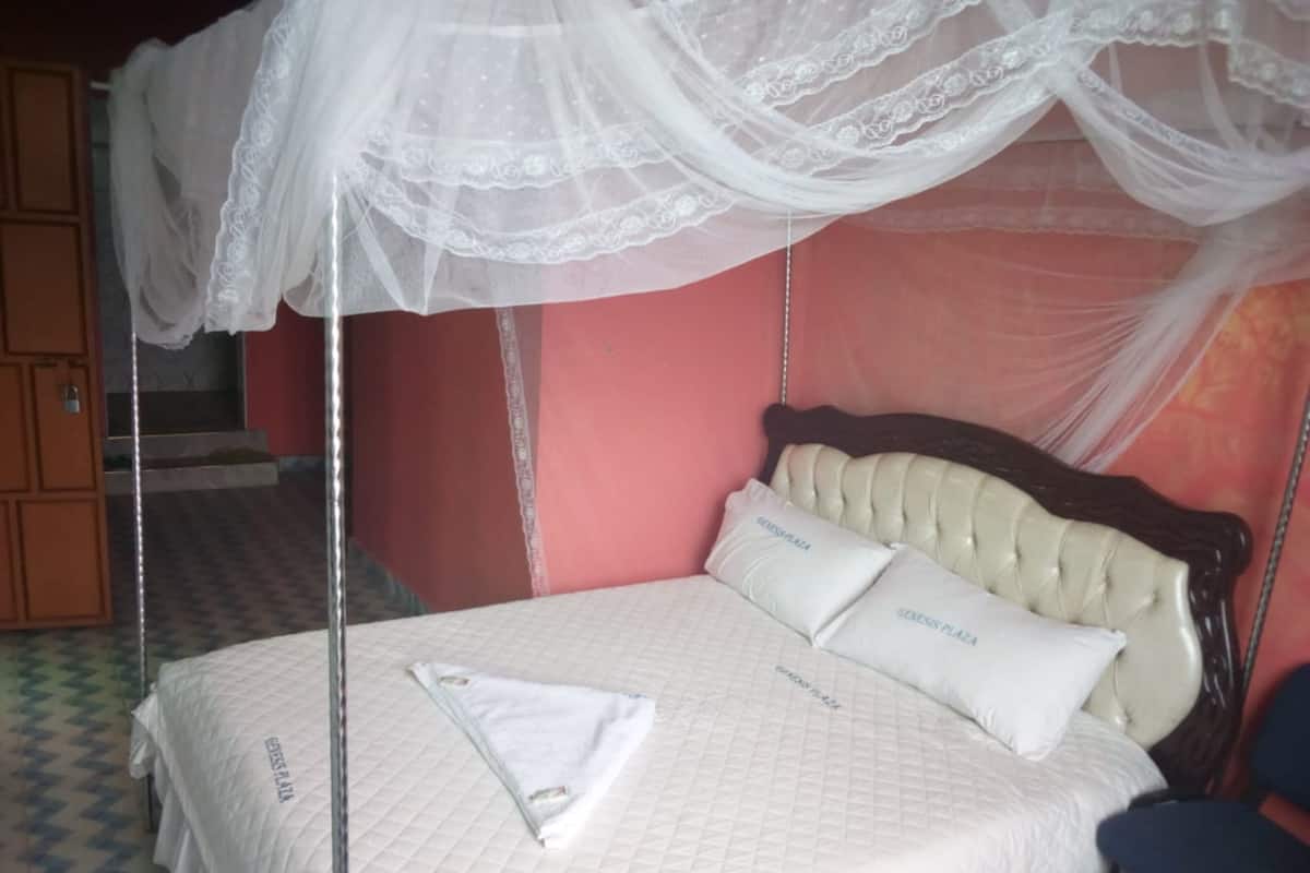 Una cama con dosel, un almohadón con el texto "GENESIS PLAZA" y una colcha acolchada.