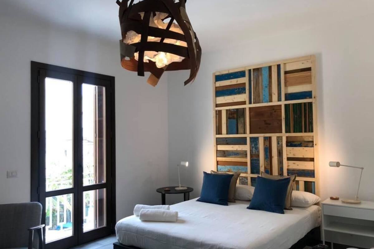 A bedroom with a large bed, a wooden wall art piece, a nightstand, and a window with a view.