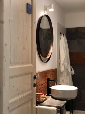 A bathroom with a wooden door, a round mirror, a white sink, and a towel hanging on the wall. - B&B Terra di Sicilia Luxury Rooms (Avola)
