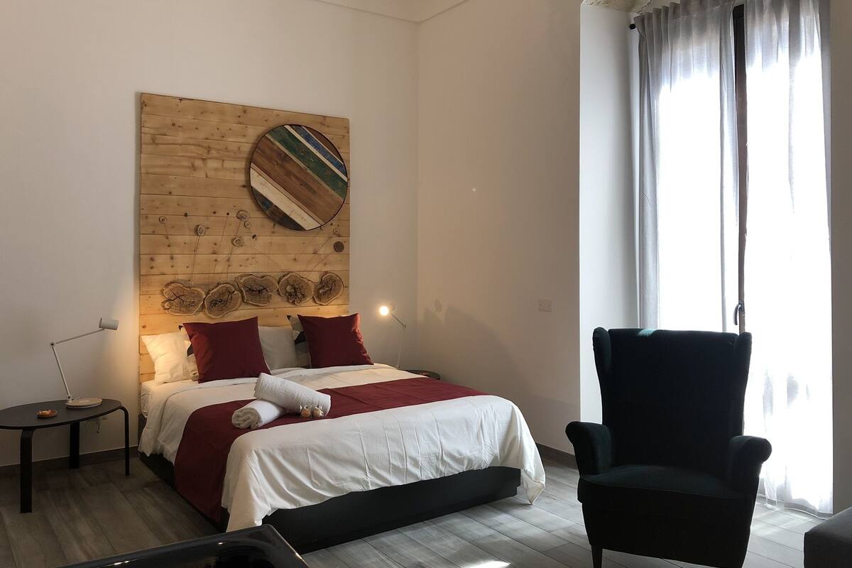 A bedroom with a bed, a chair, a bedside table, and a wall with a wooden panel and artwork.