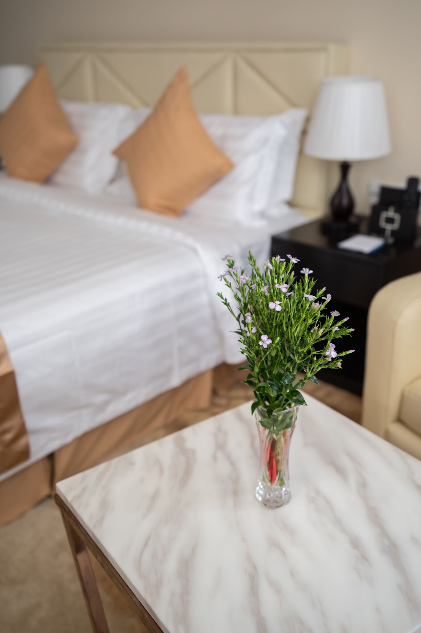 Une chambre d’hôtel avec un lit soigneusement fait, une table de chevet avec une lampe et un vase rempli de fleurs posé sur une table en marbre.