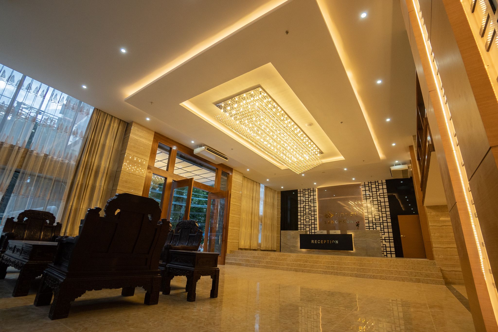 A modern hotel lobby with a reception desk, wooden furniture, and a large chandelier.