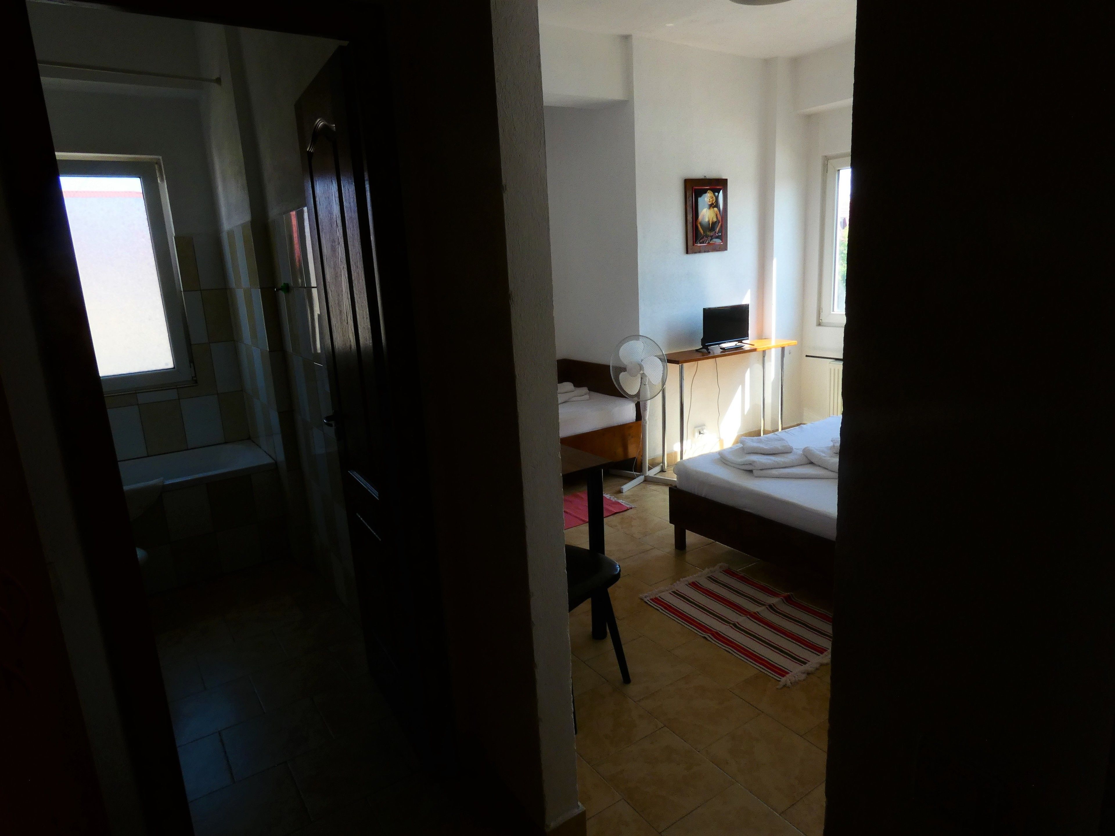 Quarto de hotel com cama, escrivaninha, computador, cadeira, ventilador e janela.