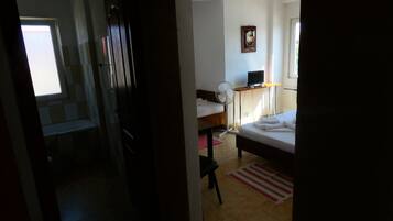 Quarto de hotel com cama, escrivaninha, computador, cadeira, ventilador e janela.