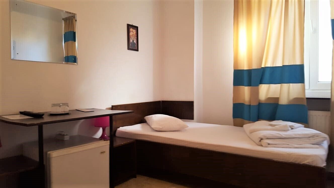 A small, single-bed room with a desk, a mirror, a small refrigerator, and a window with curtains.