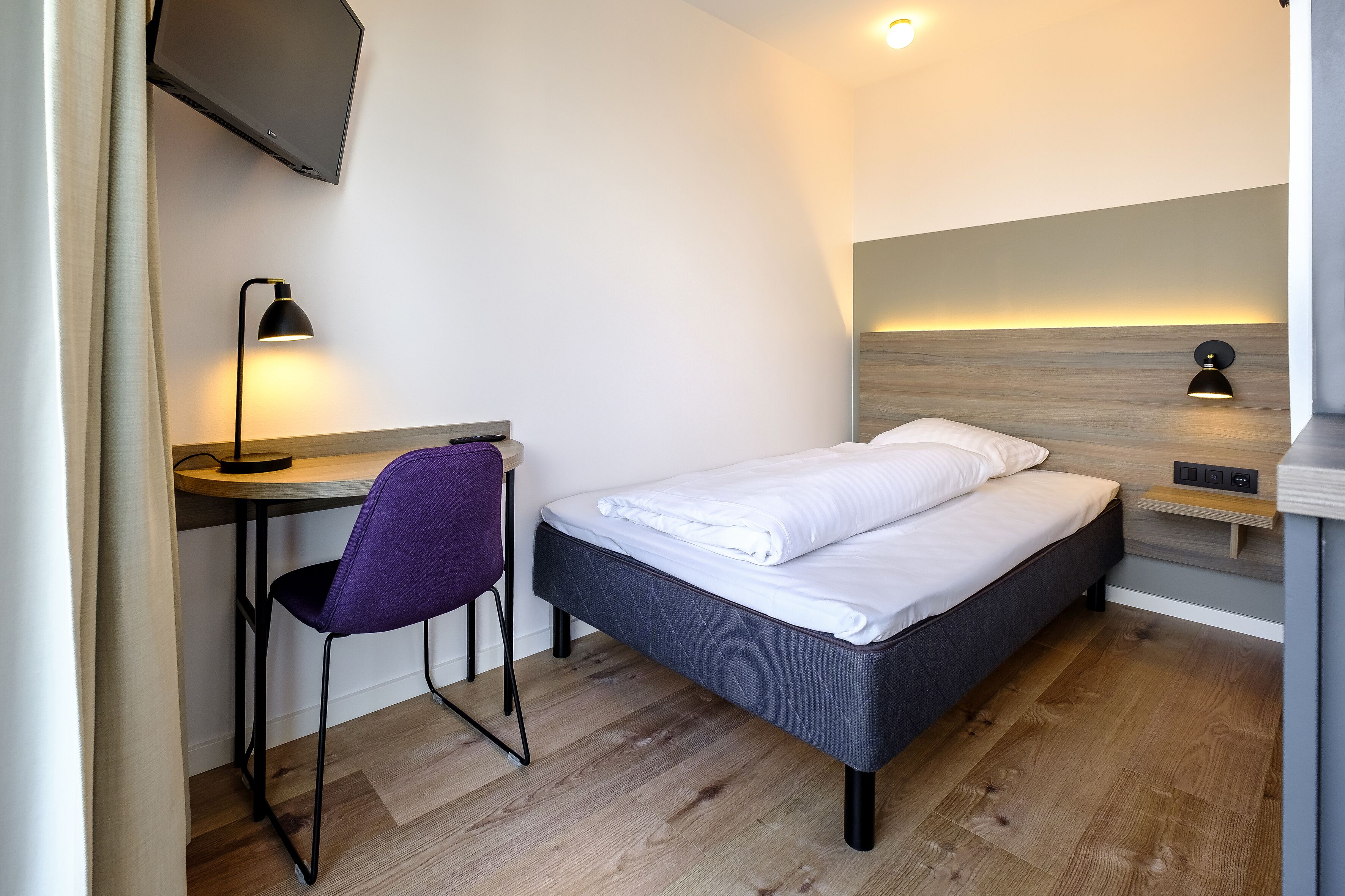 A modern bedroom with a bed, desk, chair, and two wall-mounted lamps.