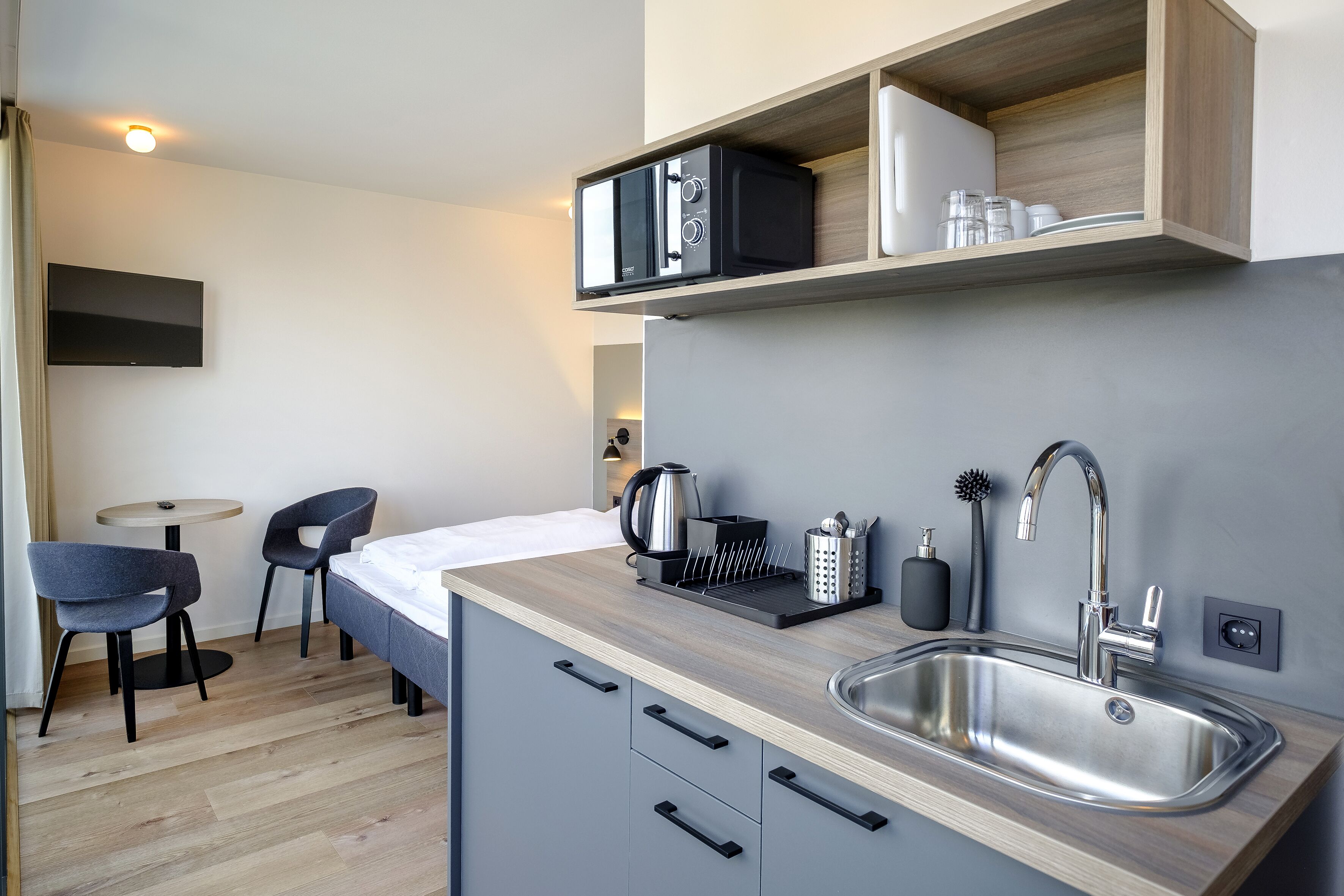Een compacte keuken met een spoelbak, koffiezetapparaat en waterkoker op het aanrecht. In het aangrenzende zithoek staan twee stoelen en een kleine tafel.