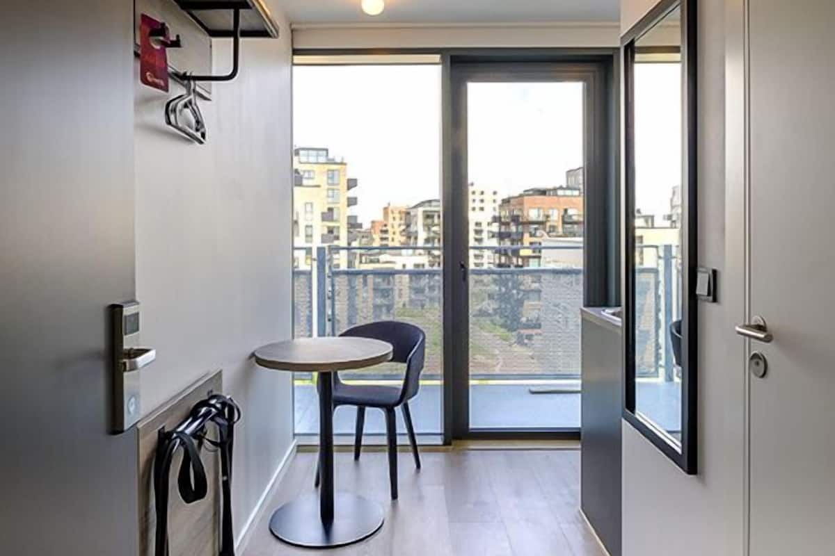 Ein schmaler Korridor mit einem kleinen Tisch und Stühlen, einer Tür zu einem Balkon mit Blick auf die Stadt und einer Deckenleuchte.