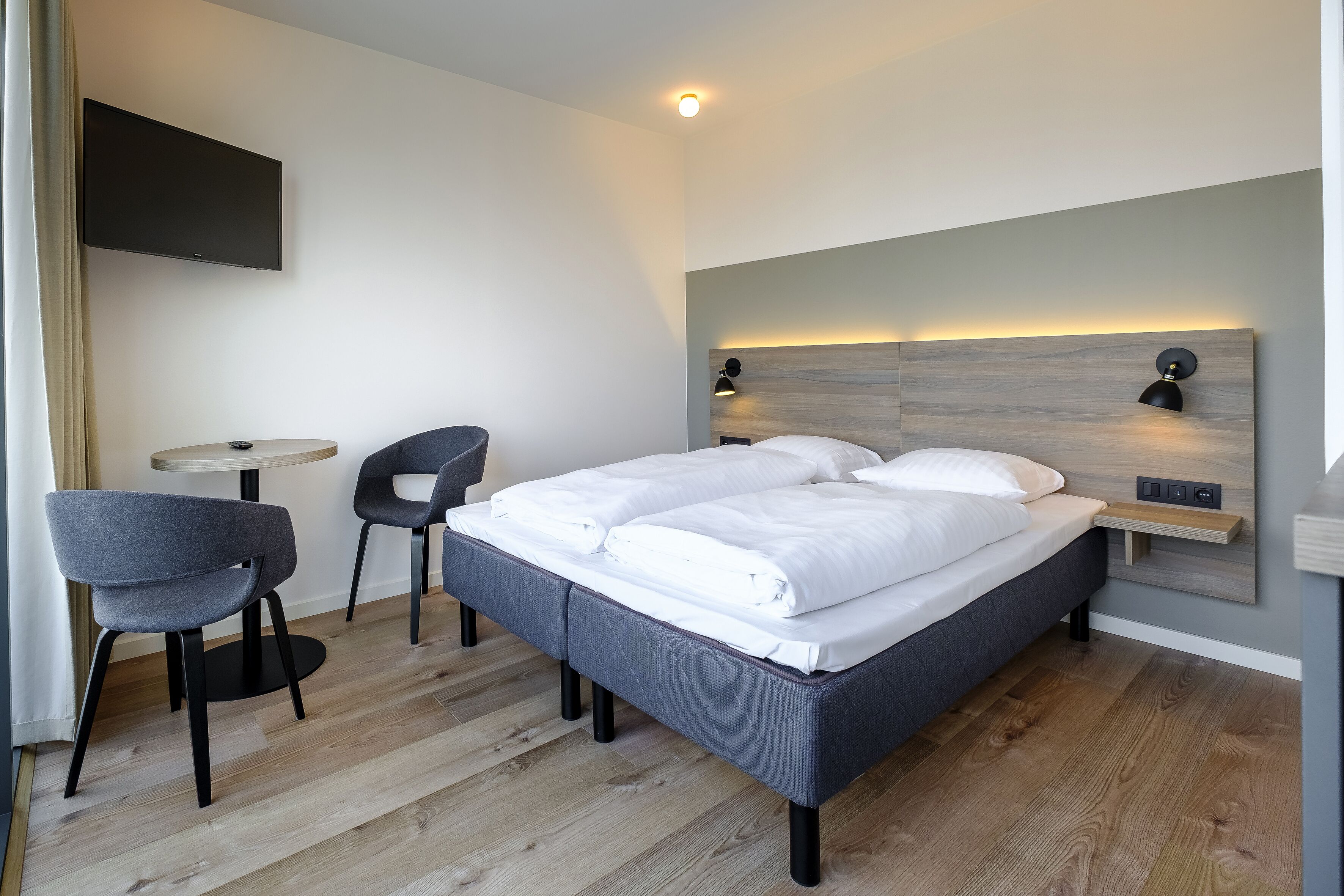 Una habitación de hotel moderna con una cama, dos sillas, una mesita y un televisor montado.