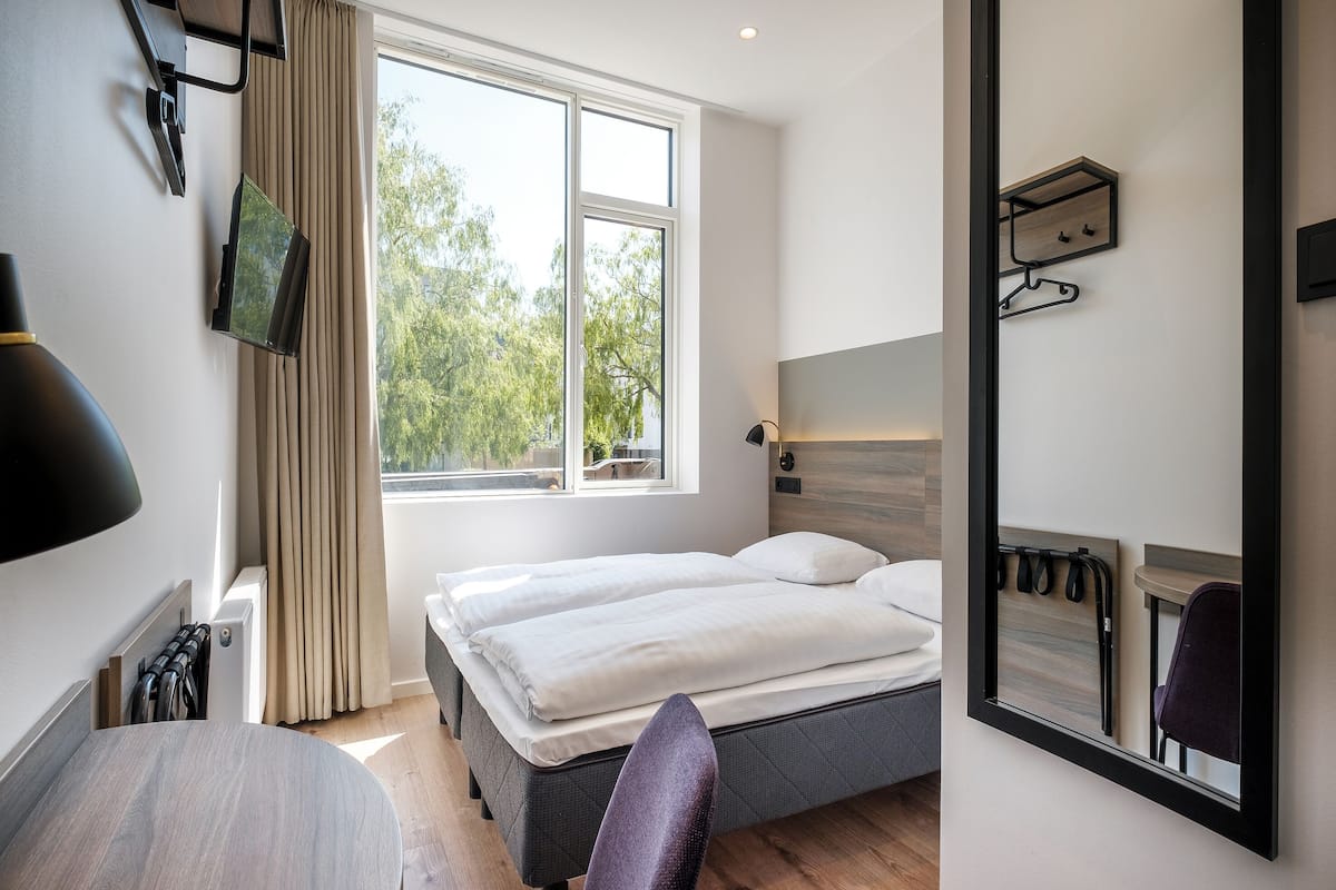 Ein modernes Hotelzimmer mit Bett, Schreibtisch, Stuhl, einem Fenster mit Blick auf Bäume und einem Spiegel.