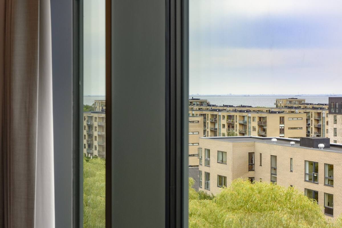 Blick aus einem Fenster auf ein Wohngebiet mit mehreren Mehrfamilienhäusern und weiter entfernten Gewässern.