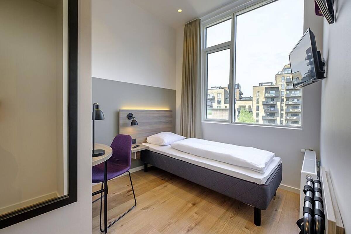 Ein kleines Hotelzimmer mit einem Einzelbett, einem Schreibtisch, einem Stuhl und einem Fenster mit Blick auf die Stadt.