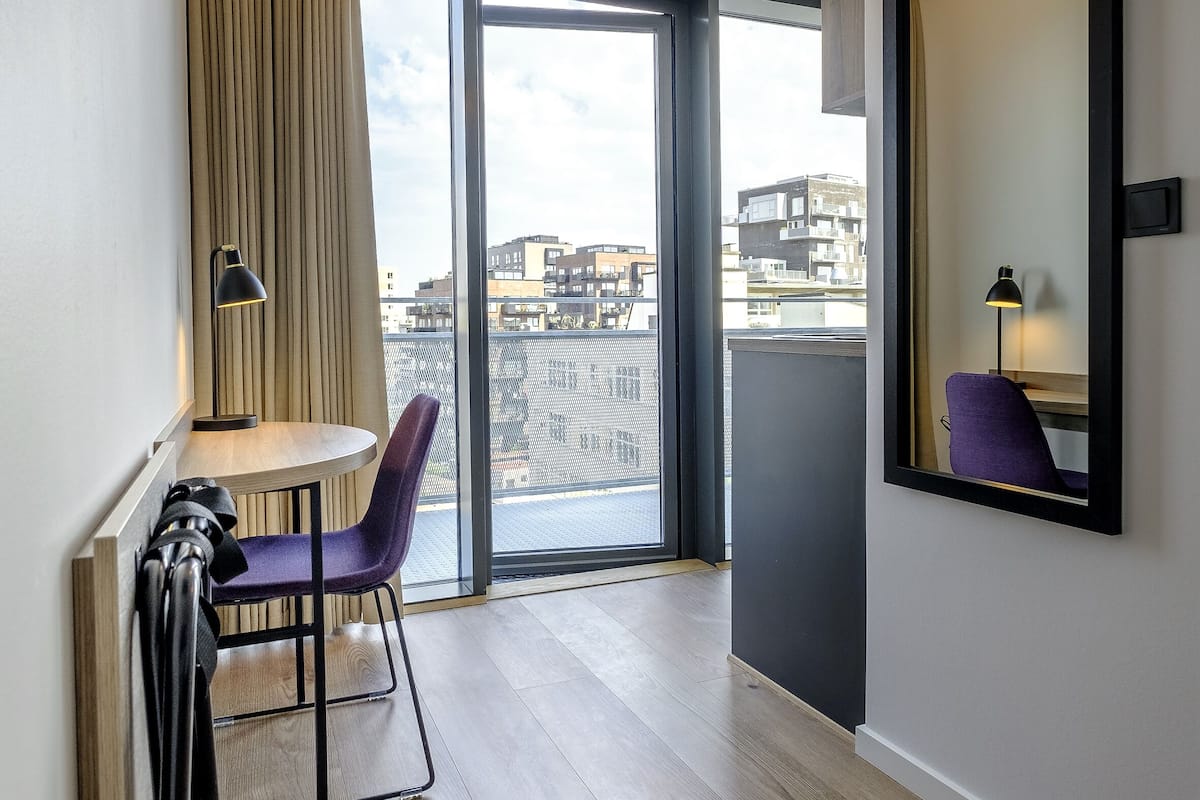 Ein modernes Hotelzimmer mit großem Fenster, einem kleinen Tisch, einem violetten Sessel und einem Spiegel an der Wand.