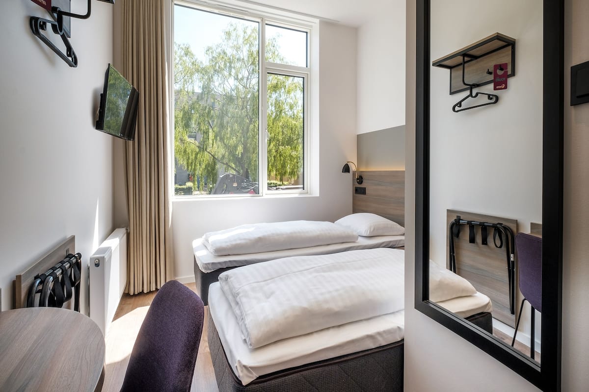 Ein Hotelzimmer mit zwei Betten, einem Fernseher, einem Fenster mit Blick auf Bäume und einem Spiegel.