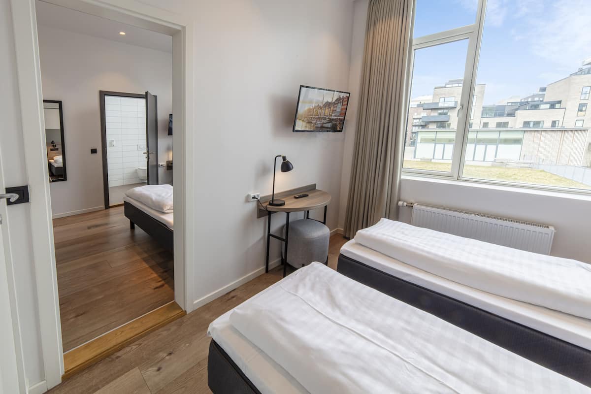 Ein Hotelzimmer mit zwei Betten, einem kleinen Schreibtisch mit Lampe, einem Bild an der Wand und einem Fenster mit Blick auf Gebäude.