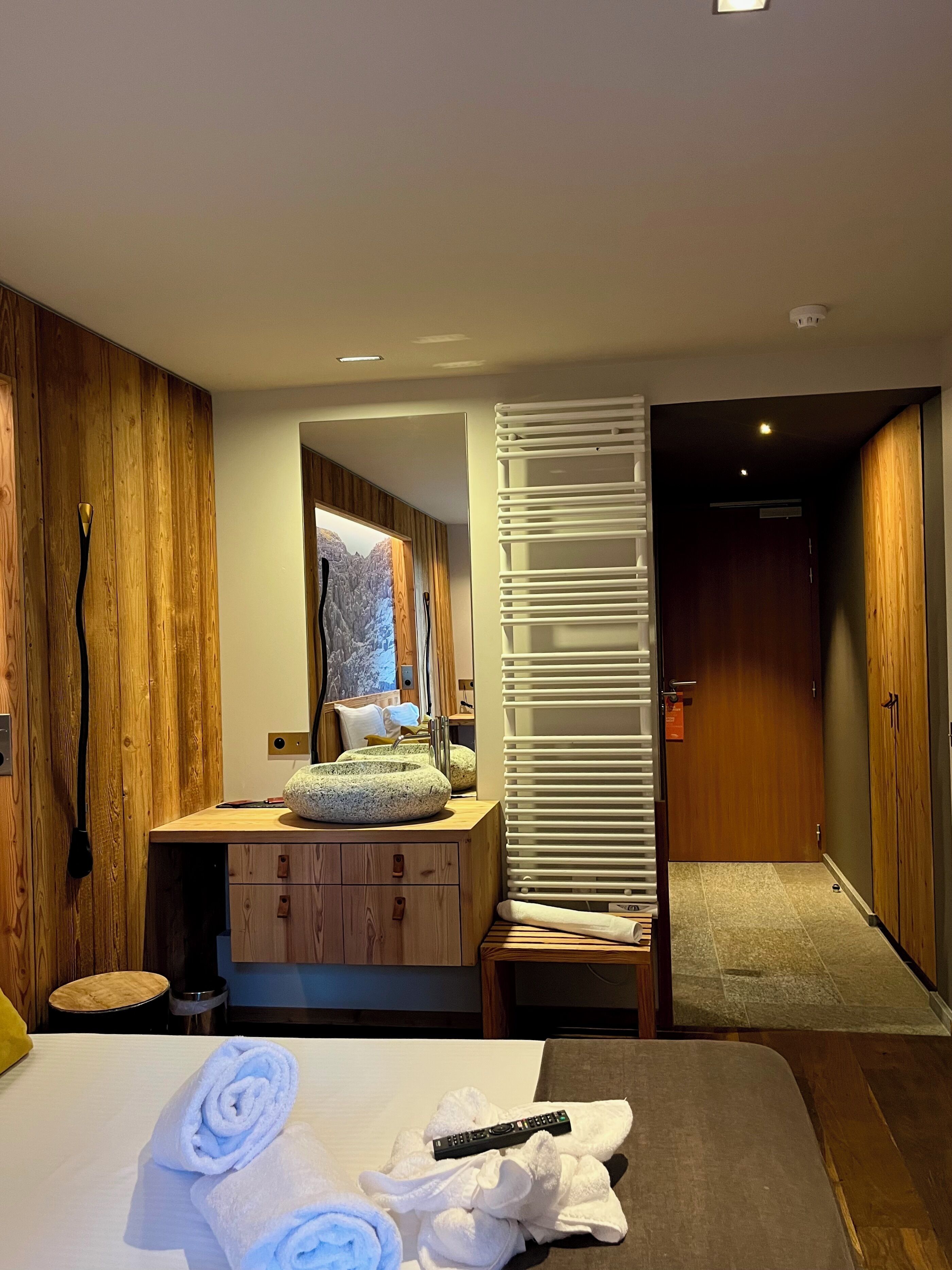Ein Hotelzimmer mit Holzwand, Schreibtisch aus Holz, einem Bett mit Handtüchern, einer Fernbedienung und einem Spiegel.