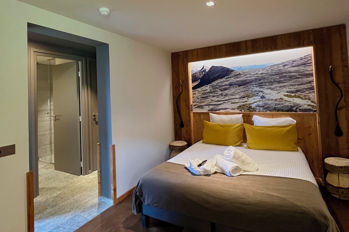 A bedroom with a bed, wooden headboard, a painting of a mountainous landscape, and a door leading to another room.