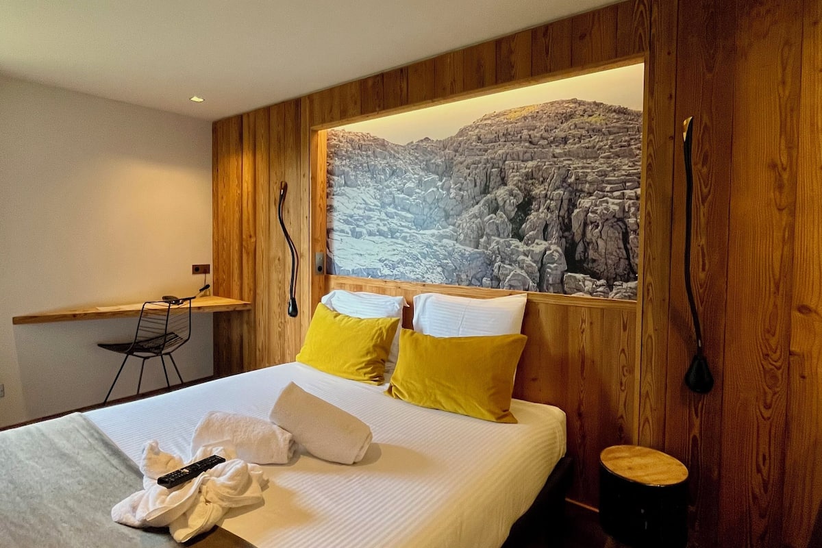 A modern hotel room with a large bed, wooden panel walls, a view of a rocky landscape, a small desk, and a chair.