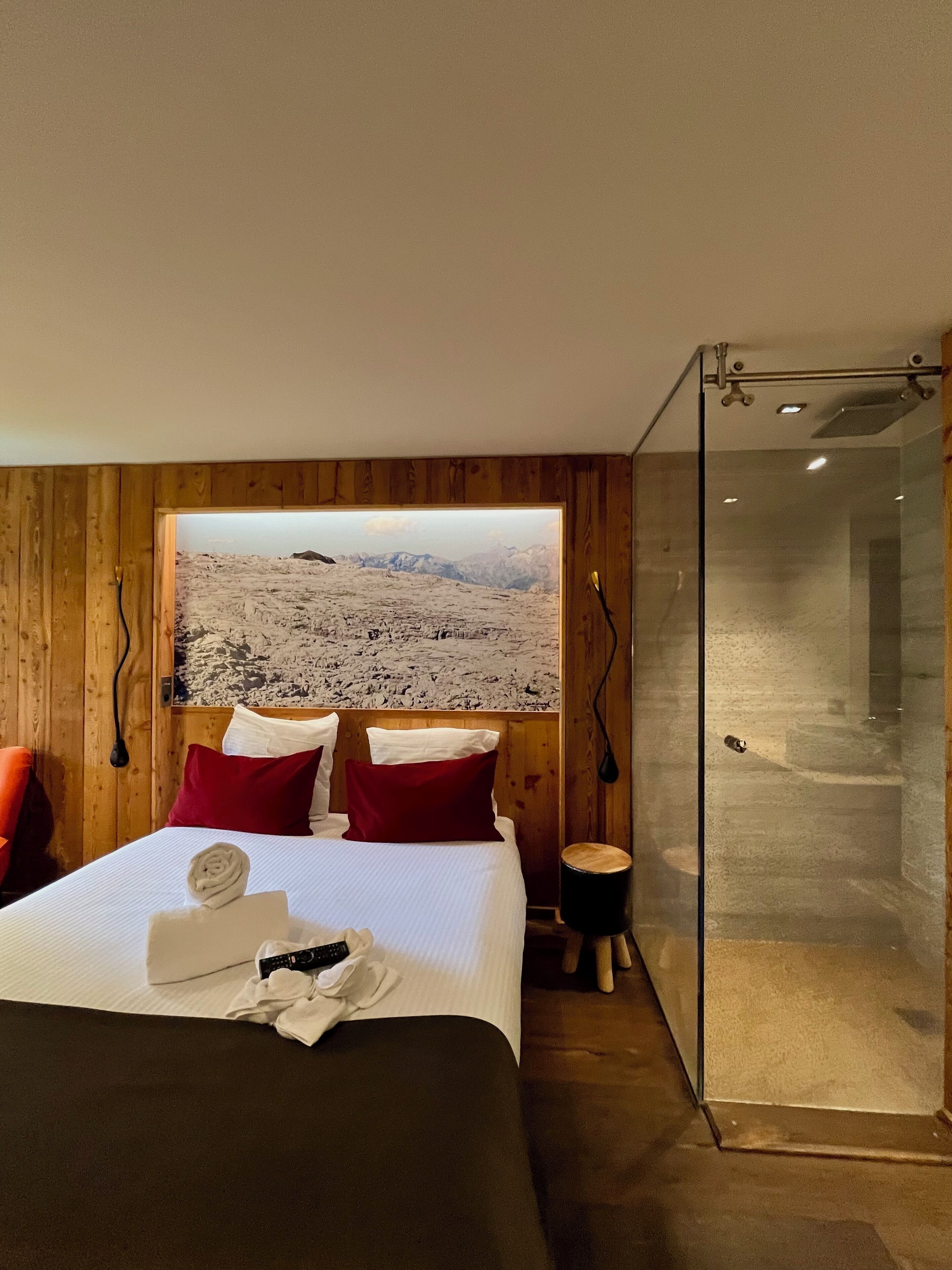 A hotel room with a large bed, a wooden headboard, a glass shower enclosure, and a view of a snowy landscape.