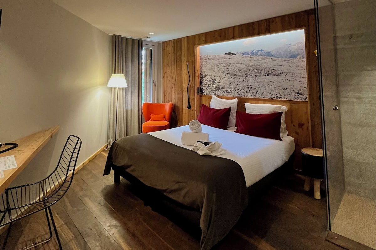A modern hotel room with a wooden floor, a bed with white and red pillows, a desk with a chair, and a large window with a mountain view.