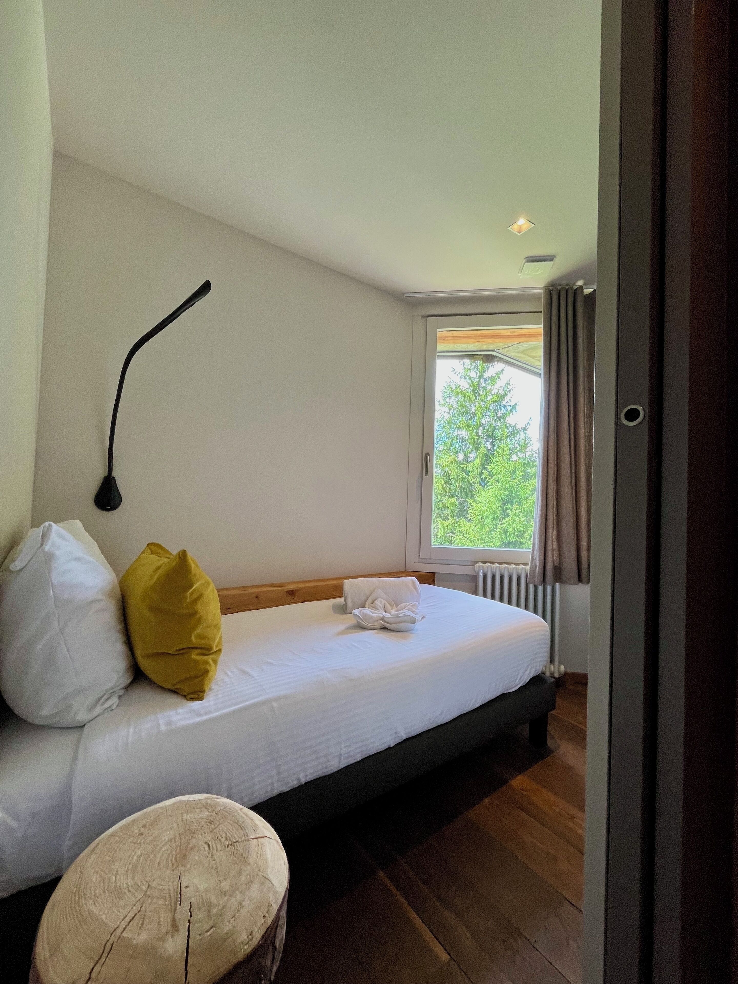 A bedroom with a bed, a bedside lamp, a window with curtains, and a view of greenery outside.