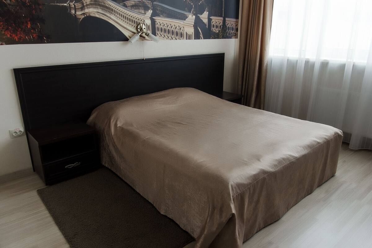 A neatly made bed with a dark headboard, a small drawer, and a dark rug on a light wooden floor.
