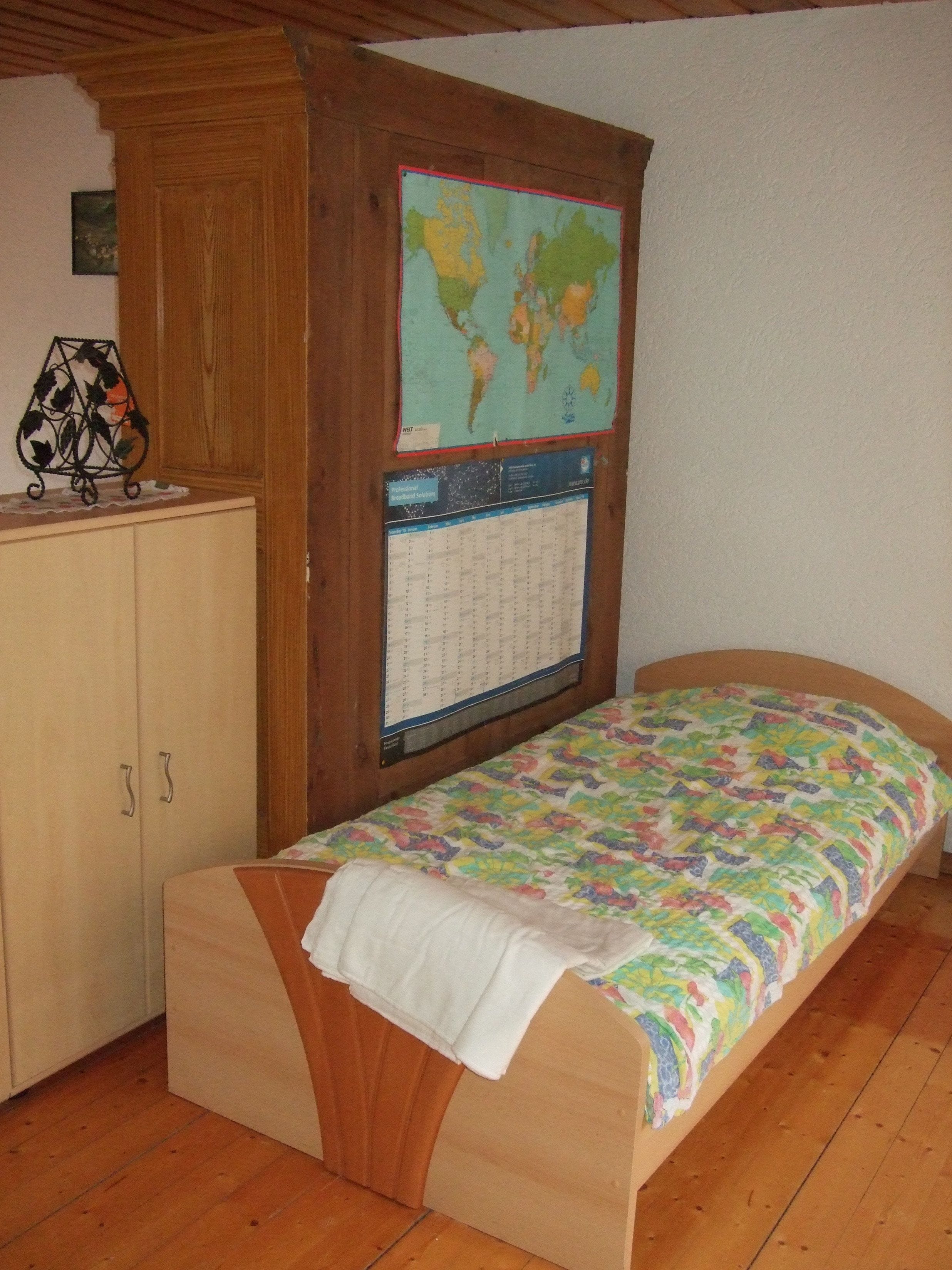 Una habitación pequeña con cama individual, cabecera de madera, un mapa mundial y un calendario en la pared.