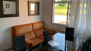 Sala de estar com sofá marrom, mesa de centro de vidro e mesa de jantar com cadeiras. Dois espelhos emoldurados na parede e uma janela com cortinas leves.