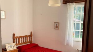 Um quarto com duas camas de solteiro, cabeceira de madeira, mesa de cabeceira com porta-retrato, luminária pendente e janela com cortinas.
