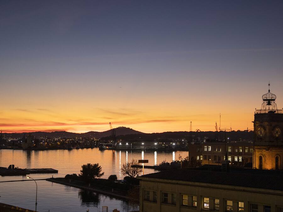 Un paisaje urbano al atardecer, con una imponente torre del reloj, agua tranquila que refleja el cielo y una colina distante.