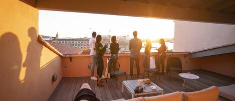 A rooftop terrace with a view of the city, wooden furniture, and a table with a hat and sunglasses.