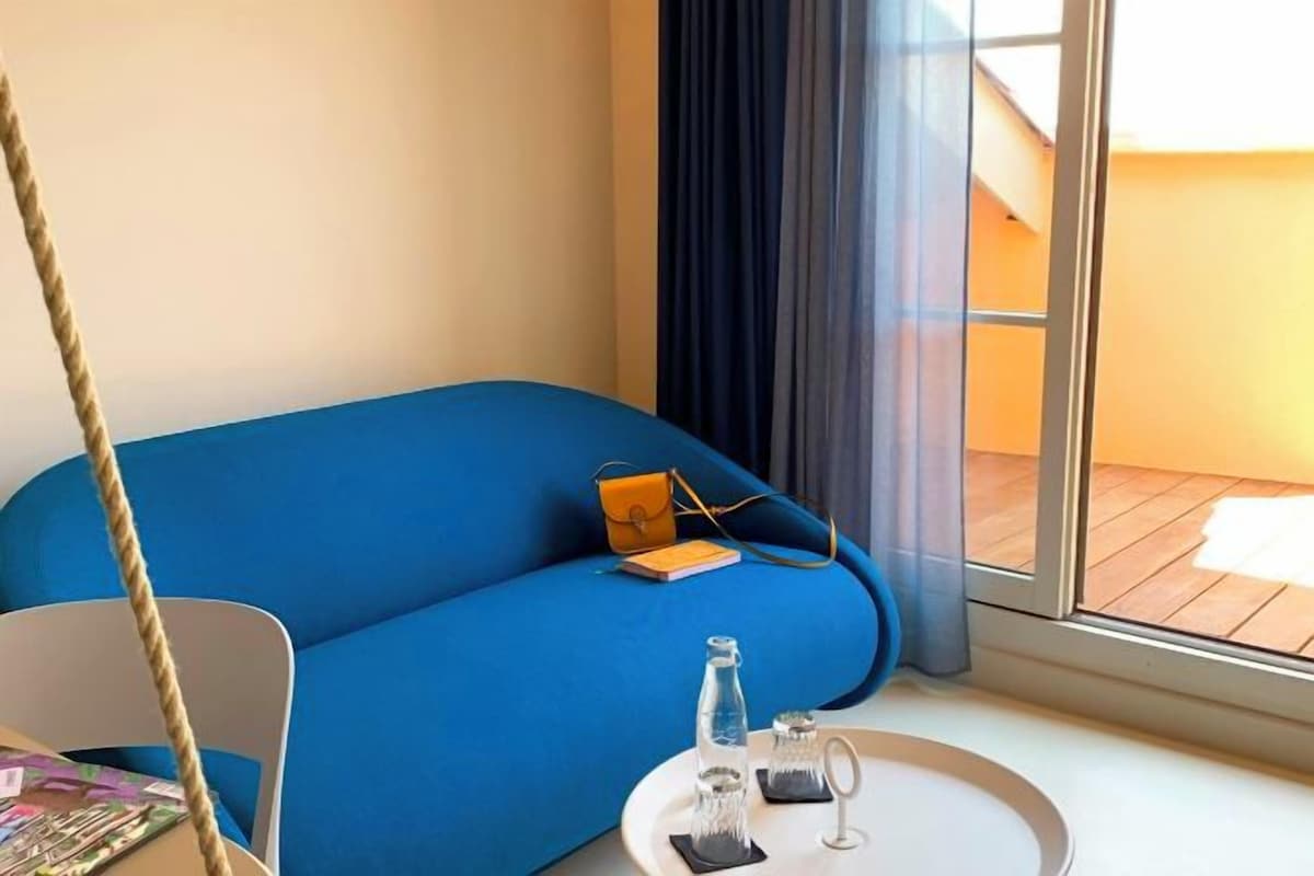 A room with a blue sofa, a small table with a bottle and glasses, a chair, and a view through a window with curtains.