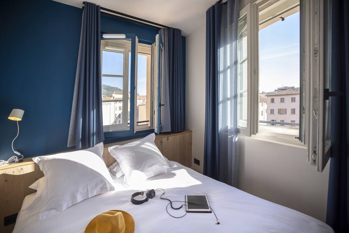 Een hotelkamer met een bed, nachtkastje met lamp, raam met gordijnen en uitzicht op gebouwen.