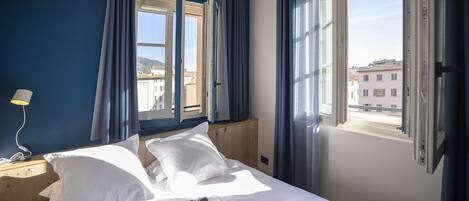 A hotel room with a bed, a bedside table with a lamp, a window with curtains, and a view of buildings outside.