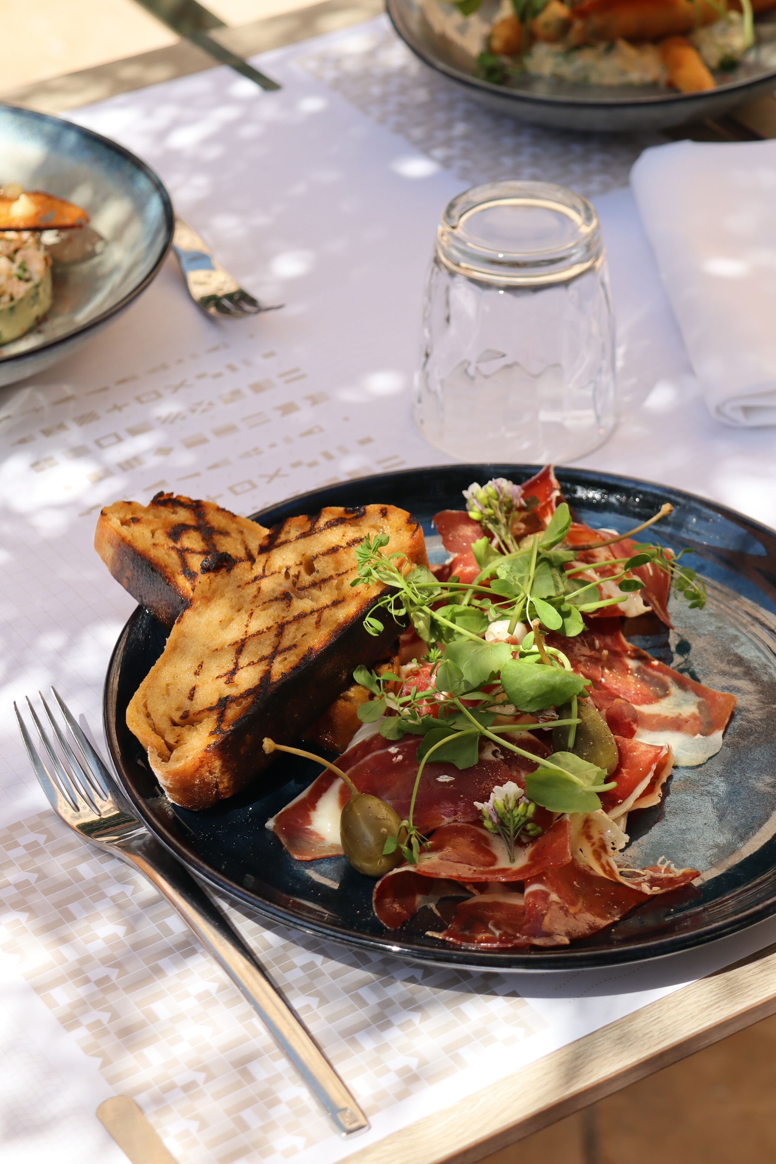 A plate of food with grilled bread, prosciutto, and greens on a table with a white tablecloth.