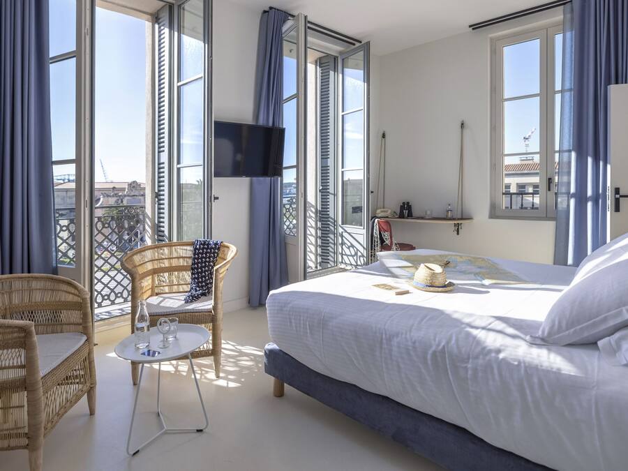 Quarto de hotel com uma cama, uma cadeira de vime, uma mesinha e uma varanda com vista.