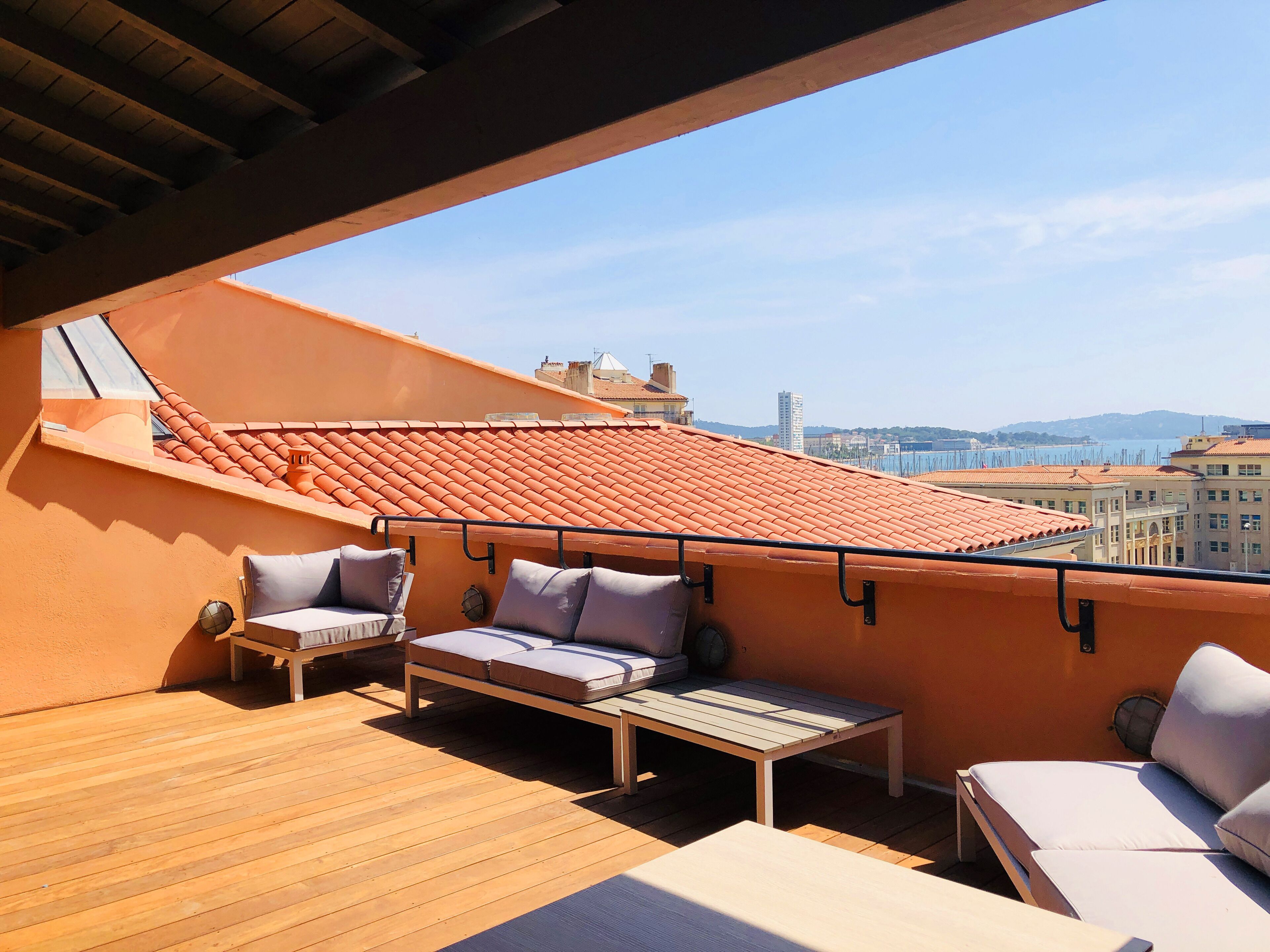 A rooftop terrace with a wooden deck, two lounge chairs, a small table, and a view of the city and sea.